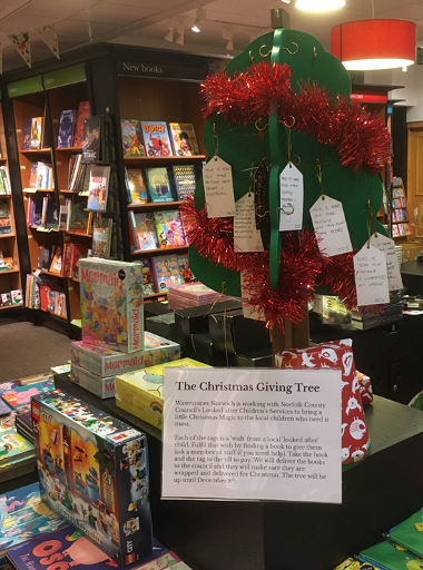 Photograph of the wooden 'Giving Tree' from 2023 in Waterstones Norwich. It is painted green with tags hanging from hooks.