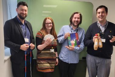 Photograph of four Multiply maths course tutors. They hold hands-on equipment such as skittles and playing cards