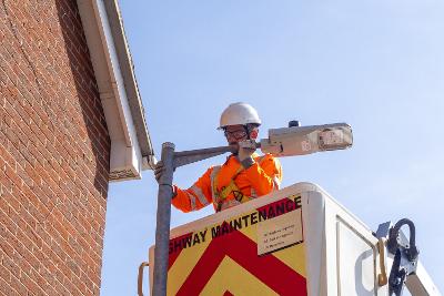 Photograph of a worker next to a streetlight. The equipment they stand on reads 'Highway Maintenance'